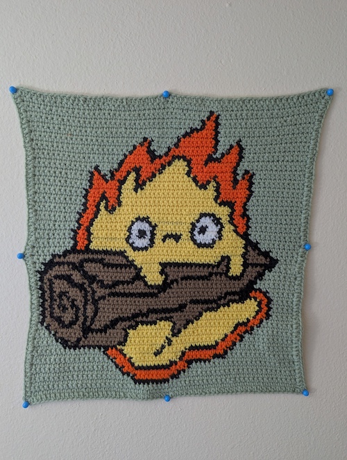 A crocheted wall tapestry depicting Calcifer resting on a log