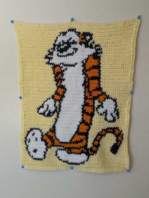 A crocheted wall tapestry depicting Hobbes on a pale yellow field