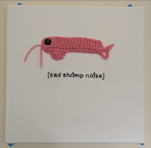 A small pink crocheted shrimp mounted on white canvas above the words sad shrimp noise